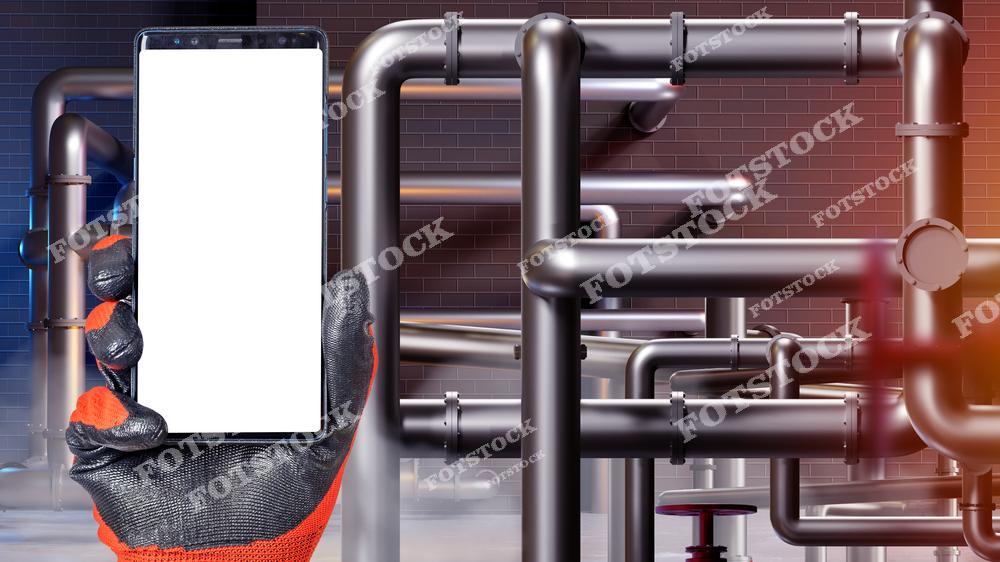 Рабочая рука в перчатке держит смартфон с белым экраном на фоне металлических труб, проходящих в разных направлениях. Фон - сложная сеть труб на стене фабрики с теплыми оттенками освещения.